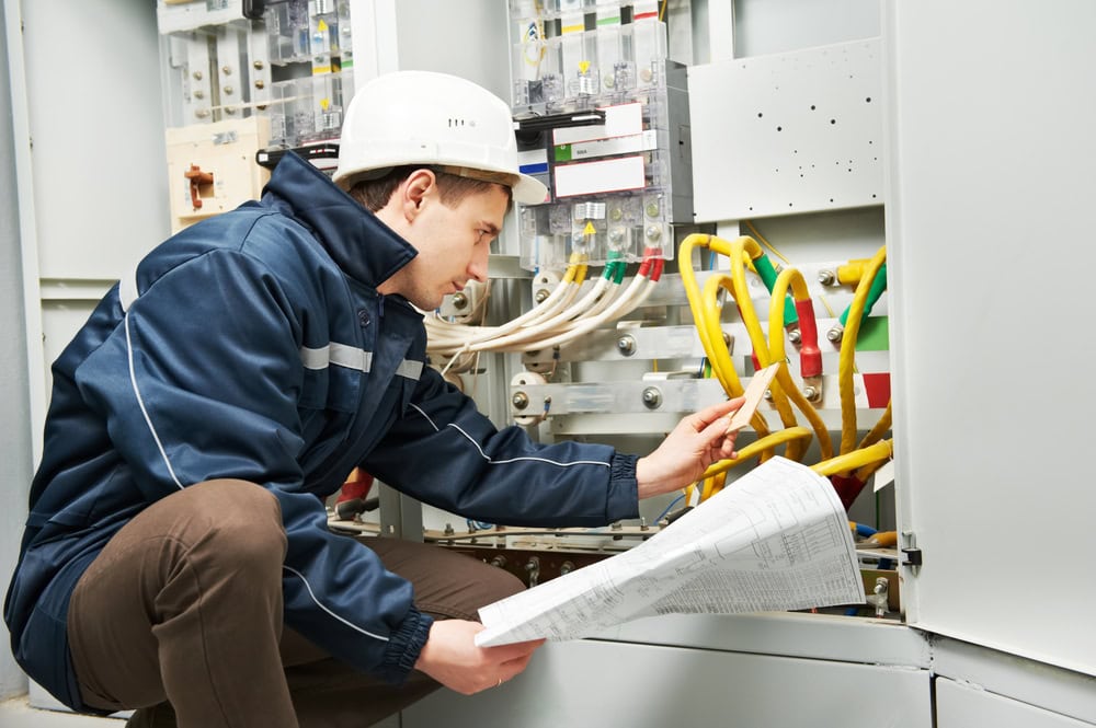 A person wearing a white hard hat and blue work jacket is kneeling while inspecting electrical equipment inside a panel. They are holding a paper document with schematics and appear to be examining the wiring and components closely, performing Electrical Services in Taylor County TX.