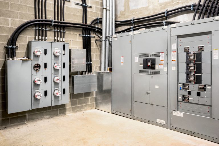 An industrial electrical room with various electrical panels and meters mounted on a cinder block wall. Conduit pipes and cables run into the panels, suggesting a complex electrical distribution system by an Electrical Contractor in Taylor County. The floor is concrete, and the environment looks clean and organized.