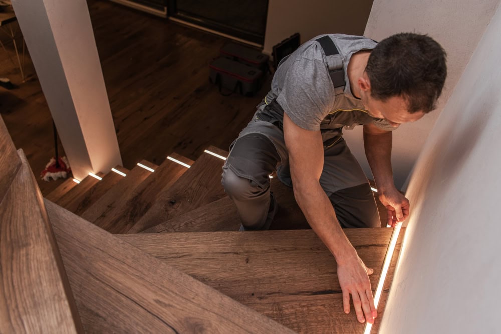 A person wearing overalls installs LED strip lights on the side of a wooden staircase. The indoor setting, with ambient lighting highlighting the steps, showcases meticulous work often seen from skilled electrical contractors in Taylor County TX. The person is focused on aligning the lights accurately along the staircase.