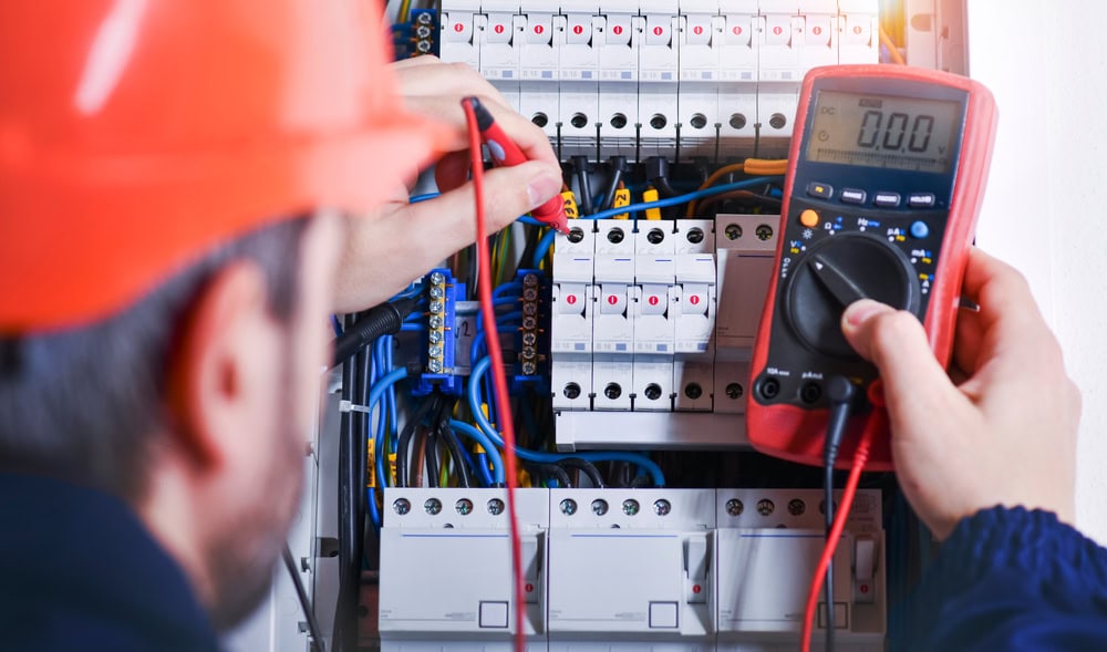 An electrician wearing an orange hard hat uses a multimeter to test an electrical panel with multiple circuit breakers and colorful wiring. The focus is on the multimeter screen showing a reading and the electrician's hands meticulously working, typical of top Electrical Contractors in Taylor County TX.