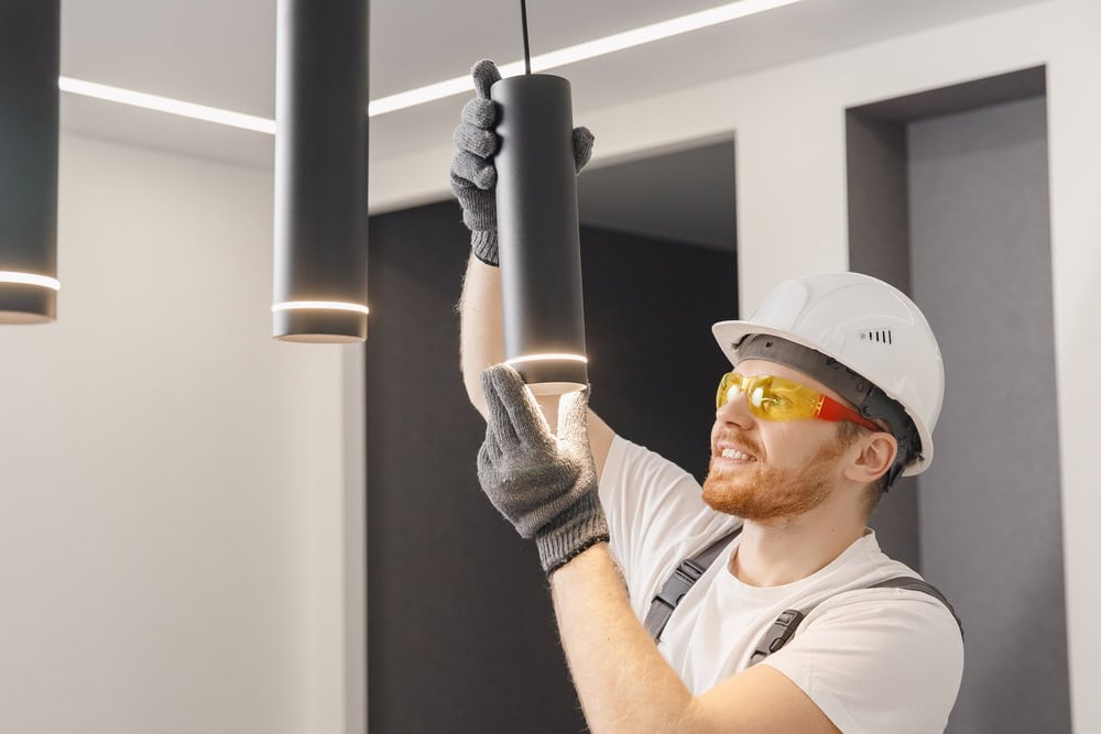 A construction worker wearing a white hard hat, yellow safety glasses, gray gloves, and a white t-shirt is adjusting a black cylindrical ceiling light fixture in a modern indoor space. This Taylor County TX electrician is meticulous with lighting installation to ensure top safety standards.