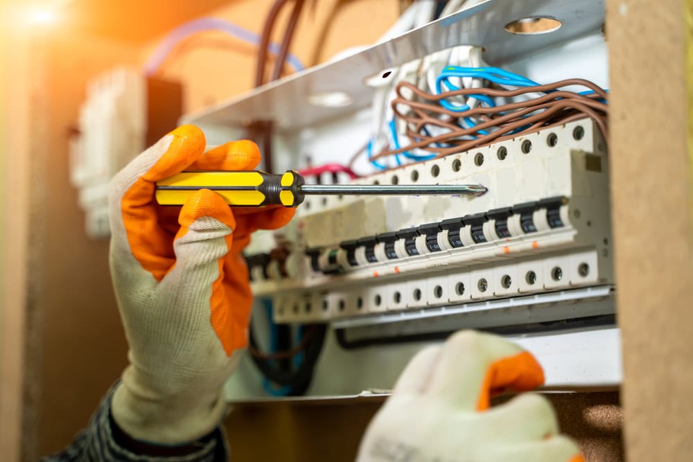 A person wearing gloves uses a yellow and black screwdriver to adjust components in an electrical panel. The panel contains various wires and circuit breakers. The background is softly lit, highlighting the electrical contractor's focused work on the system.