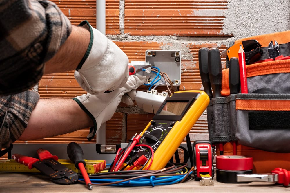 An electrician wearing gloves uses a screwdriver to work on an open electrical outlet in a brick wall. Various tools, including a multimeter, pliers, tape, and a tool bag, are placed on a nearby surface. This scene highlights the expertise of an Electrical Contractor in Taylor County.