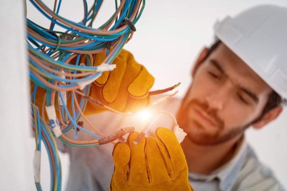 A close-up of a construction worker in a white hard hat and yellow gloves working on wiring. The worker, an electrical contractor from Taylor County, is focused on connecting several colored wires, which are slightly glowing at the connection point. The background is blurred.