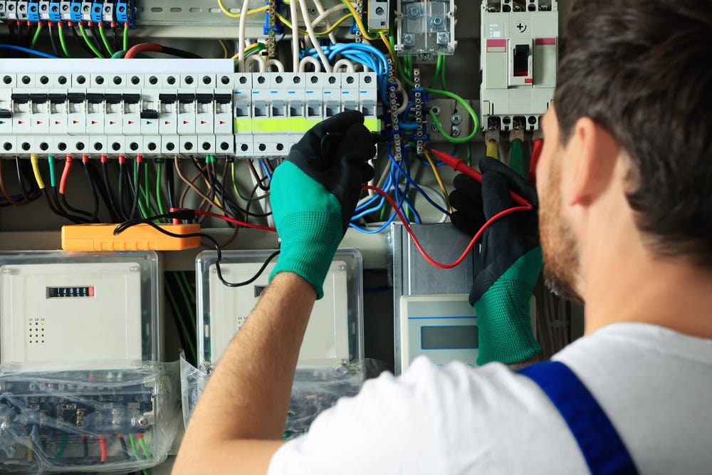 A technician wearing green gloves is working on an electrical panel, testing the connections with red and black wires. The panel is filled with various colored wires and components. As he focuses on his task, his back to the camera, it’s evident that reliable Electrical Services are crucial for safety.