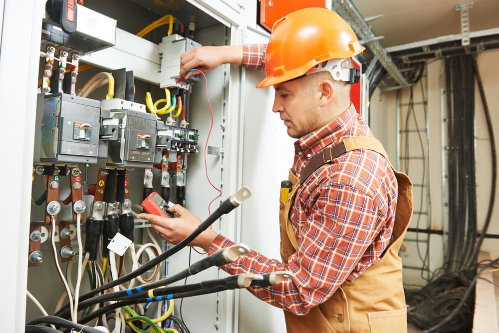 A construction worker wearing a hard hat and plaid shirt is examining an electrical panel with various wires and circuit breakers. As an experienced Electrical Contractor in Taylor County, he uses a testing tool to ensure the setup is correct and safe. The background shows additional cables and part of a building's infrastructure.