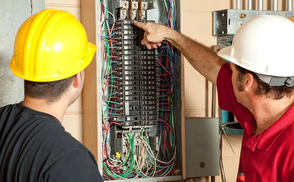 Two electricians wearing hard hats are inspecting an electrical panel in Taylor County. The person on the right, in a red shirt, is pointing at a specific circuit breaker while the person on the left observes. The panel is open, showing various wires and components, highlighting their expertise in generator installation.