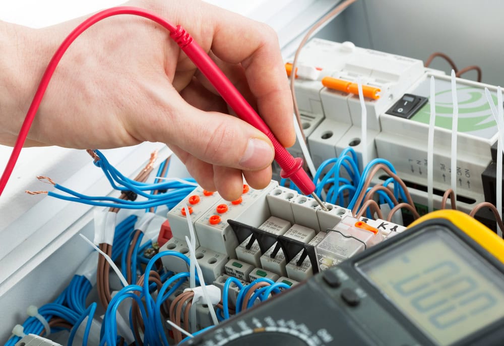 A close-up of a hand using a red probe to test electrical connections inside a control panel with various wires and components. A yellow multimeter in the foreground shows a reading of 0.00, suggesting routine diagnostics or maintenance by an Electrical Contractor in Taylor County.