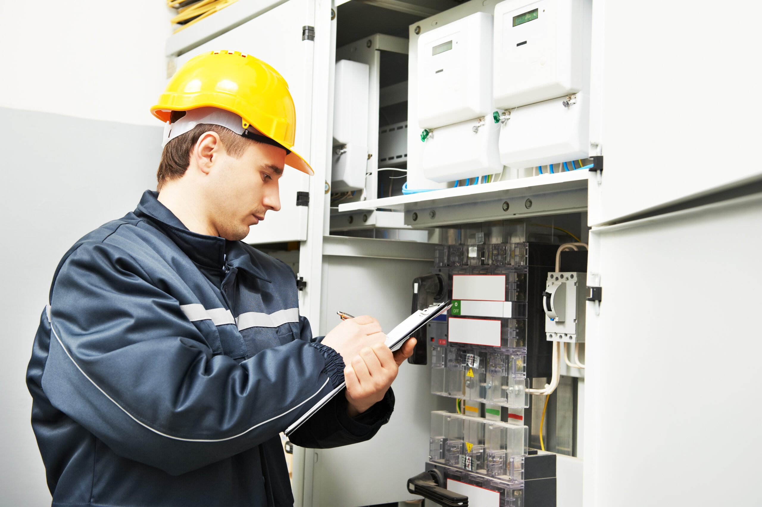 An electrician in Taylor County, wearing a yellow hard hat and a dark jacket, is inspecting an electrical panel. He holds a clipboard and seems to be taking notes. The panel is filled with various components and meters, highlighting the expertise required for tasks like generator installation in Taylor County.