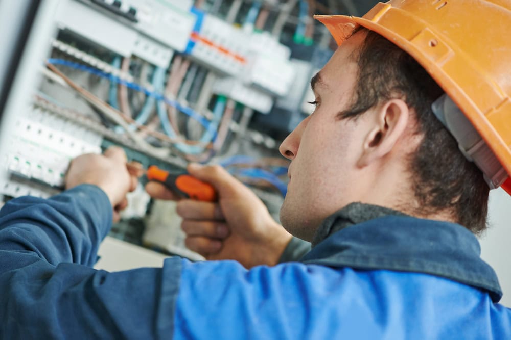 An electrician wearing an orange hard hat and blue work uniform uses a screwdriver to work on a wiring panel with several wires and circuit components, showcasing the expertise required for Generator Installation in Taylor County.