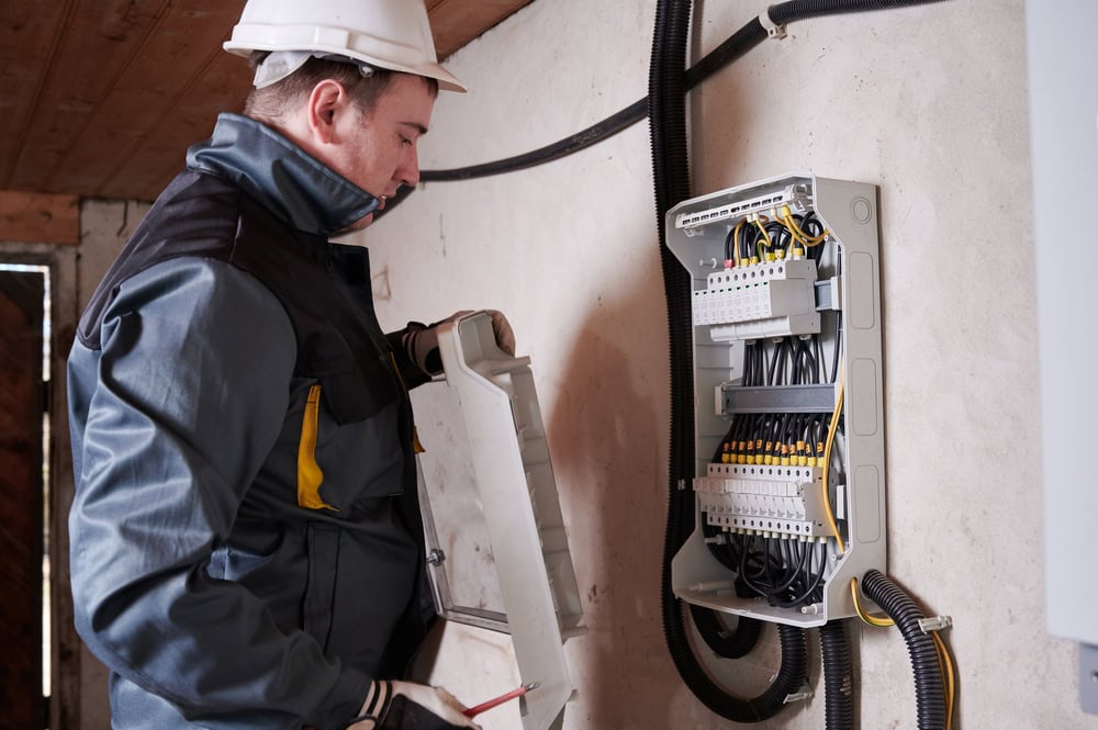 A technician wearing a white hard hat and protective clothing is inspecting an open electrical panel. He is holding the panel cover with one hand and a screwdriver with the other. The panel, part of a Generator Installation in Taylor County, is mounted on a white wall with multiple cables organized inside.