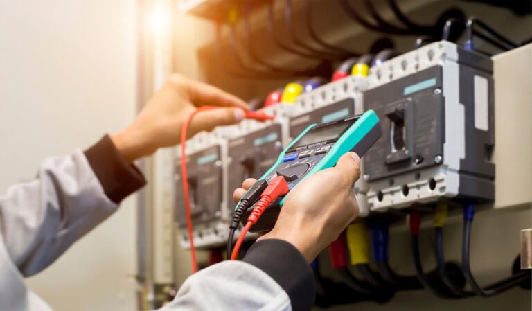 A person holds a digital multimeter to a control panel, testing electrical connections. The panel, possibly part of a generator installation in Taylor County, has various switches, knobs, and wires. The background is blurred slightly, focusing on the multimeter and the person's hands. Warm lighting illuminates the scene.