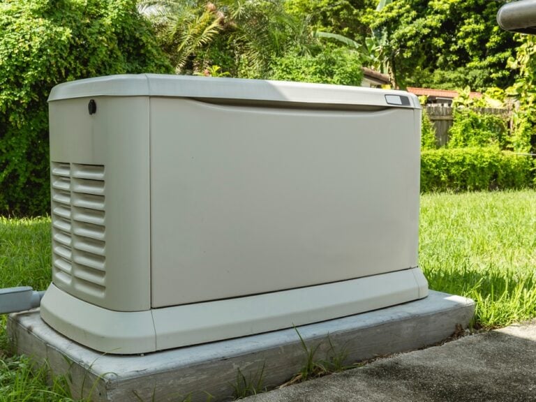 A beige generator, expertly installed on a concrete platform, sits in a grassy yard with dense greenery behind it. Its vented sides and sleek design allow it to blend seamlessly with the natural surroundings.