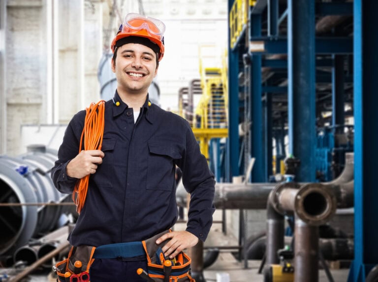 A smiling electrician in Taylor County, wearing an orange helmet, protective glasses, and navy blue overalls, holds a coiled orange cable over his shoulder. He stands in an industrial setting with machinery and pipes in the background.