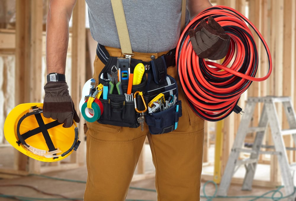 An Electrical Contractor in Taylor County, dressed in brown pants and a gray shirt with a tool belt full of tools, holds a yellow hard hat in one hand and a coiled red cable in the other. The background shows the wooden framing of a building under construction.