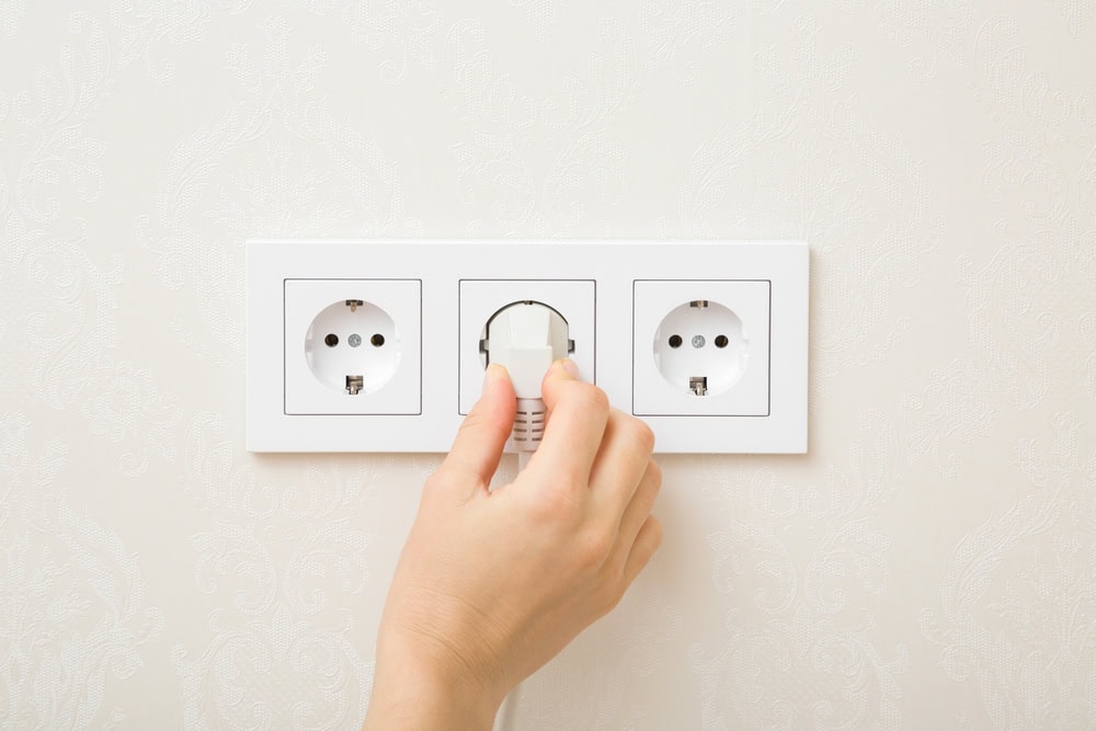 A hand is plugging a white electrical cord into the middle socket of a triple wall outlet on a textured white wall. Two adjacent sockets remain empty.