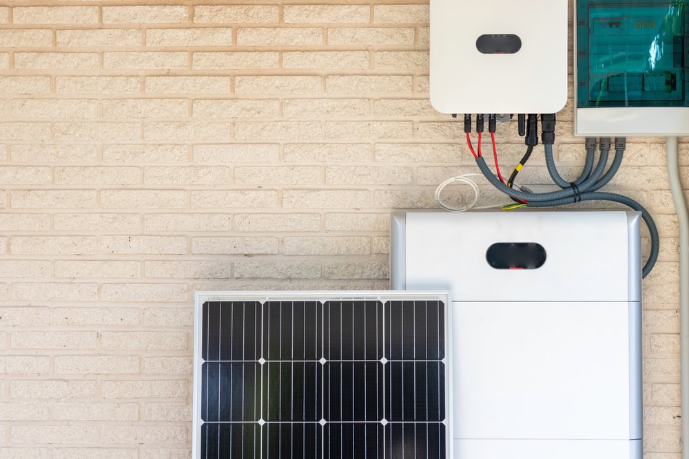 Solar power system on a brick wall with a solar inverter and battery storage unit connected by wires. A solar panel leans against the battery unit, showcasing a sustainable energy setup.