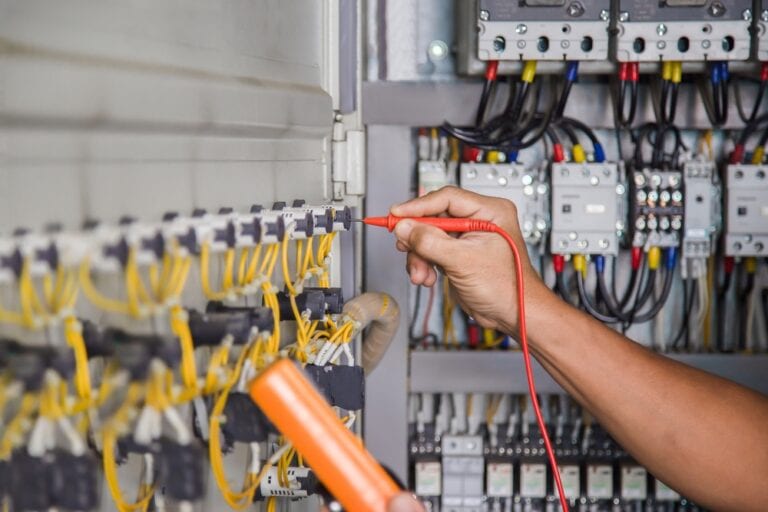 A person is using a red electrical tester to examine the connections in a circuit breaker panel filled with yellow and black wires. Various switches and relays are visible in the background.