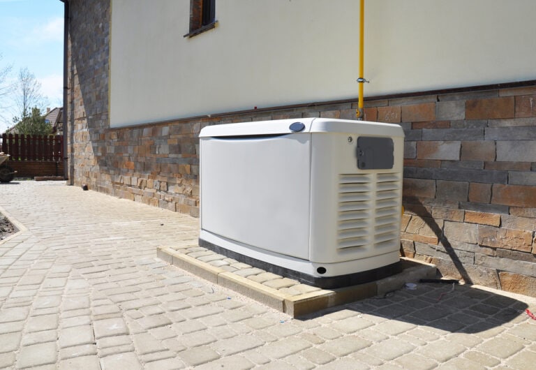 A large, white standby generator is placed on a concrete platform next to a building with stone siding. The area is paved with interlocking bricks and a yellow pipe extends from the generator upwards.