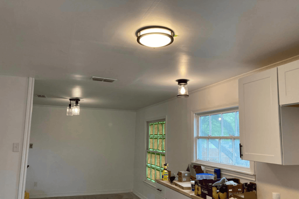 The bright room, with its white walls and ceiling, showcases the expertise of electricians in the placement of three ceiling lights. Daylight floods through a window on the right, illuminating kitchen cabinets and assorted items scattered on a countertop below.