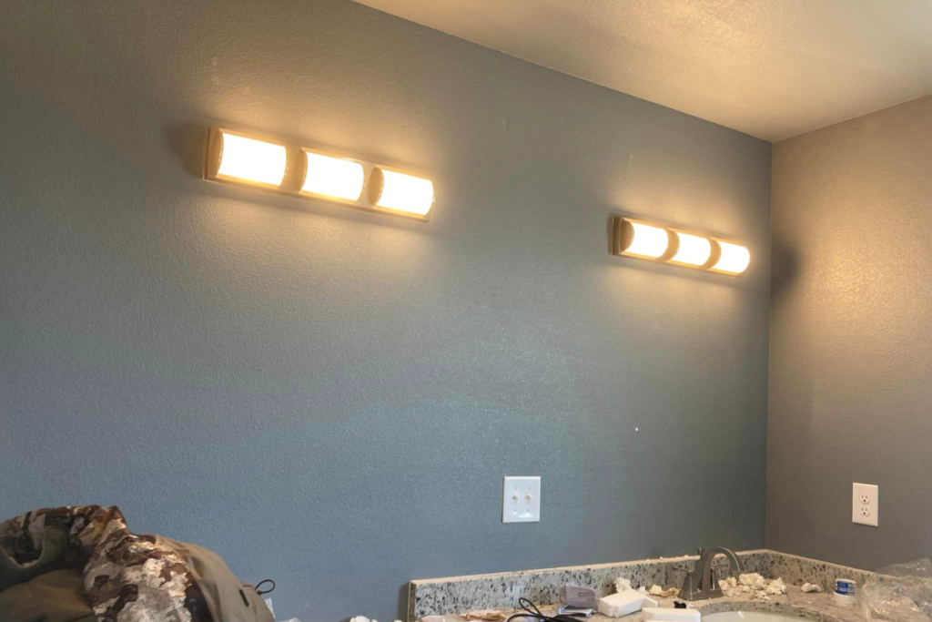 A bathroom with a gray wall features two bright, rectangular light fixtures expertly installed by electricians above a granite countertop. The countertop has some items scattered, and there's a small piece of clothing visible on the left side.