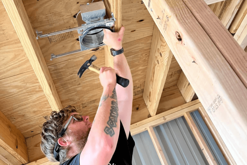 A person with glasses and a tattoo is standing on a ladder like a seasoned electrician, hammering a nail into wooden rafters as they expertly work on an electrical fixture in the building under construction.