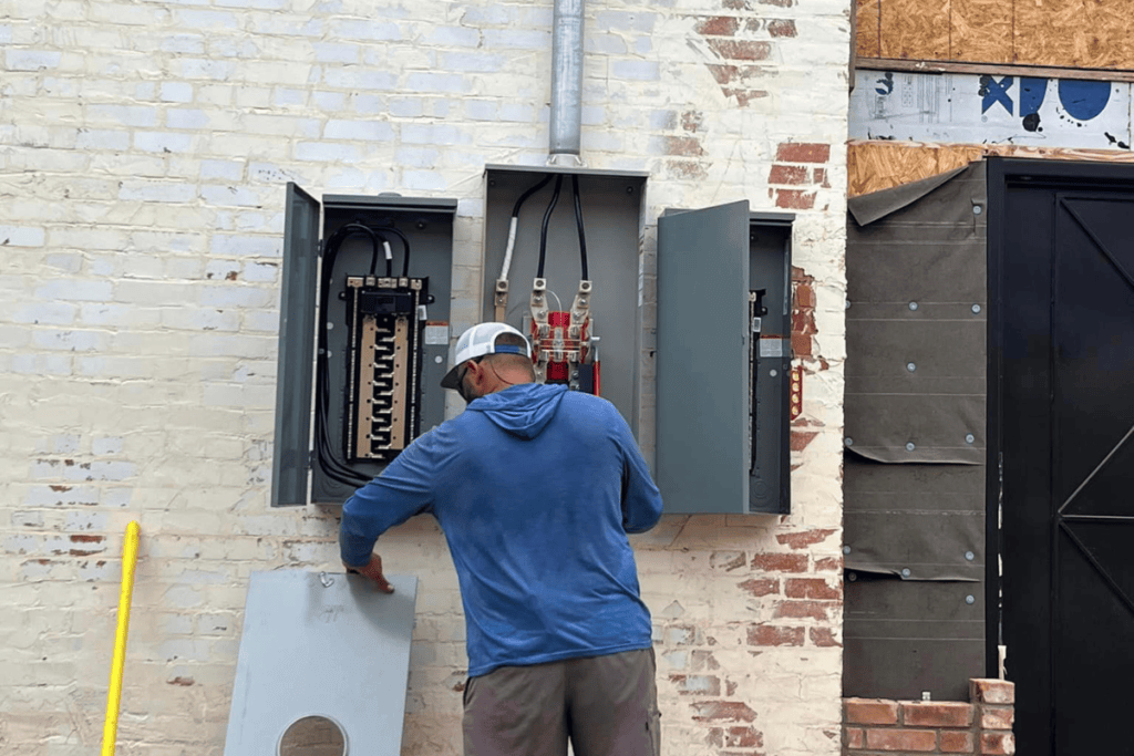 An electrician in a blue hoodie and white cap is diligently working on an electrical panel attached to a brick wall. With the panel box open, various wires and circuits are exposed. A partially boarded-up area is visible on the right, adding a sense of urgency to their task.