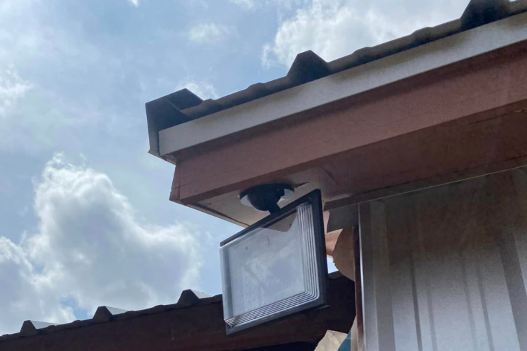 A rectangular outdoor light fixture, expertly mounted by skilled electricians, is set beneath the edge of a roof with corrugated panels. The sky is partly cloudy in the background.