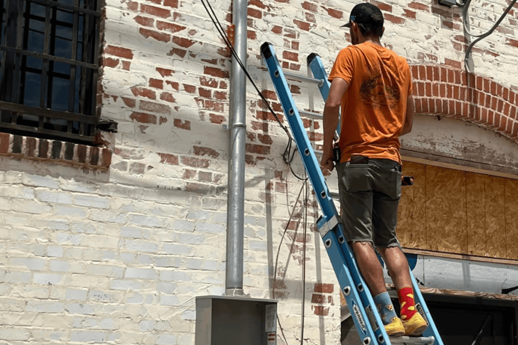 An electrician in an orange shirt, shorts, and colorful socks stands on a blue ladder, tending to a brick building. A window with metal bars is on the left, while an arched brick design crowns a boarded-up section of the wall.