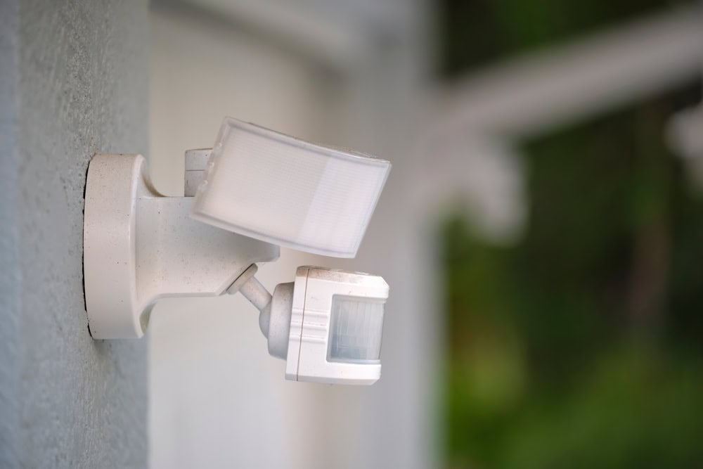 A white motion sensor light is mounted on a gray wall. The device has two main components: a light fixture on top and a sensor below. The background is blurred with shades of green, suggesting foliage or outdoor surroundings.