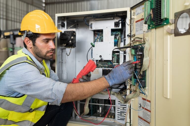 A technician wearing a yellow hard hat and safety vest is repairing an industrial machine. He is using a red multimeter to test electrical components inside an open control panel.