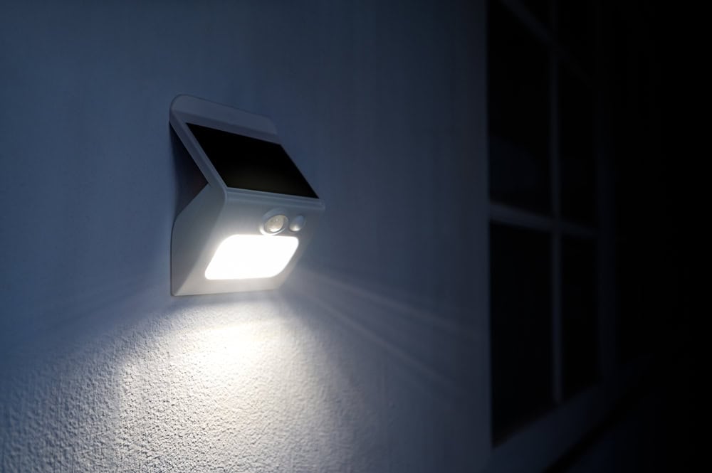 A solar-powered motion sensor light is mounted on an exterior wall, casting a bright white glow in the dark. The light design is sleek and modern, and it illuminates a nearby window frame.