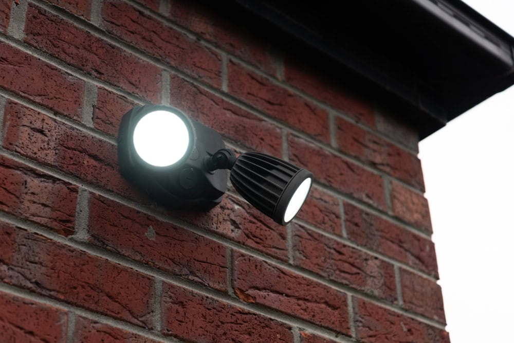 A security light mounted on a red brick wall is illuminated during daytime. The light consists of two circular lamps aimed in different directions. The sky is overcast.